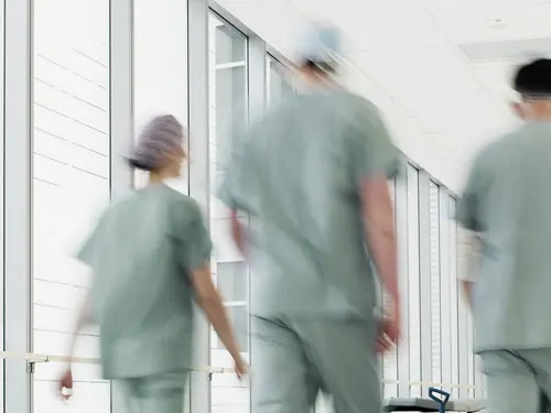 Interiør fra et sykehus, tre helsepersonell vandrer i en lang gang i grønne klær med ryggen til.