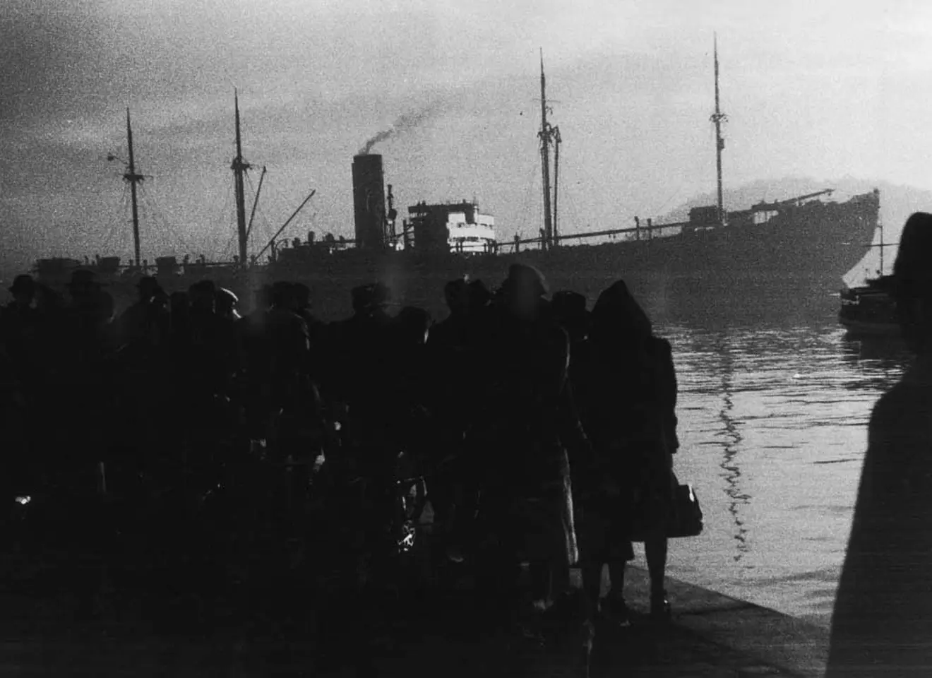 Kornete sort-hvitt fotografi med personer i forgrunnen og et skip i bakgrunnen
