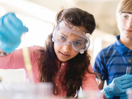 Studenter som gjør vitenskapelig eksperiment i klasserommet. Jente med blå hansker og vernebriller, gutt i bakgrunnen  med vernebriller og reagensrør