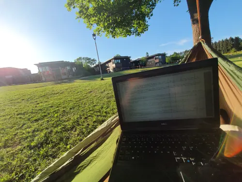 Geir Arne Hjelle i hengekøye med laptop og utsikt over solfylt gressplen og nabolag 