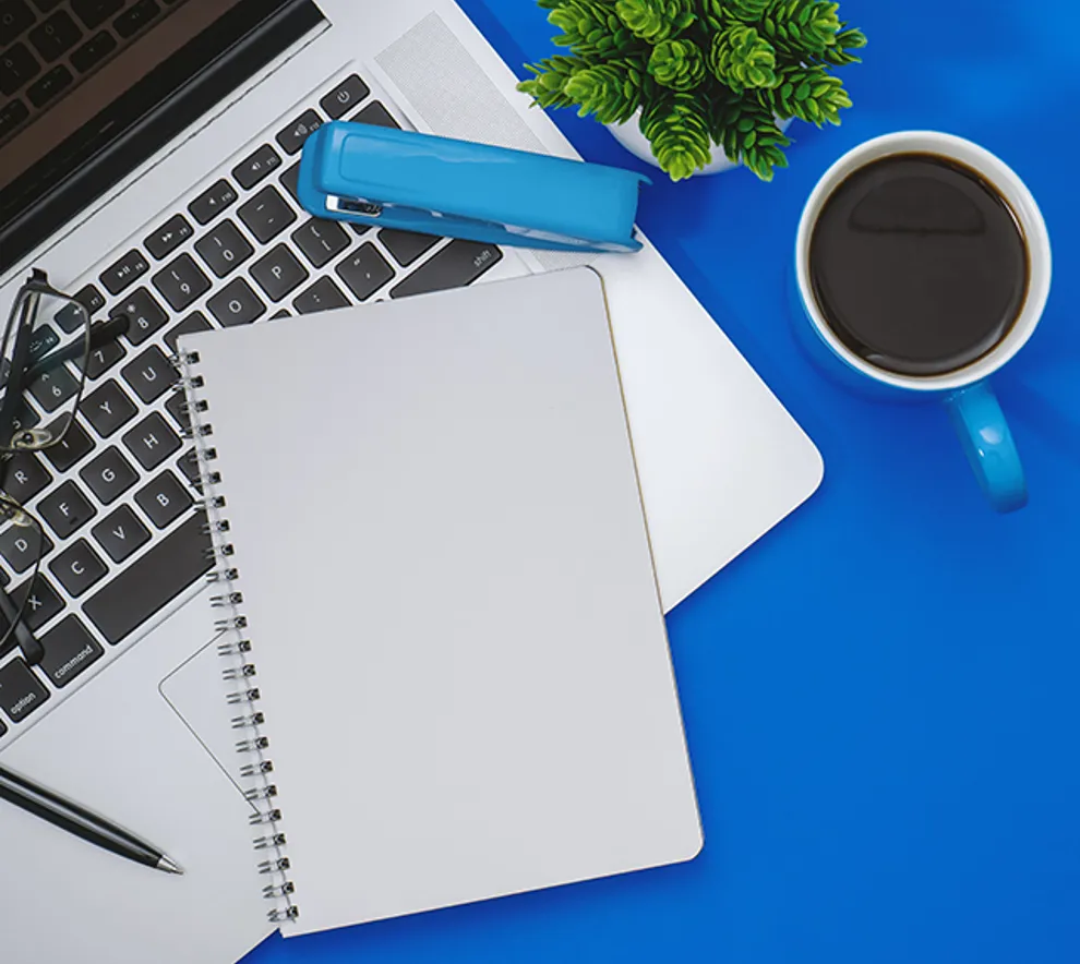 Mac, skrivesaker, briller, stiftemaskin, kaffekopp og en grønn plante på blått underlag