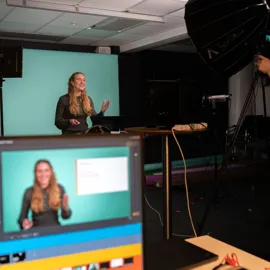 Kvinne står foran kamera og presenterer noe, lyskaster i bakgrunnen og monitor i front
