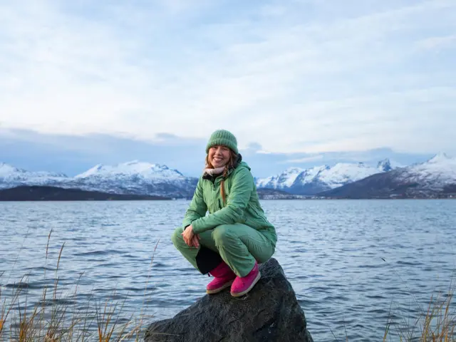 Kvinne på huk på fjæresteiner med  fjord og fjell  bak