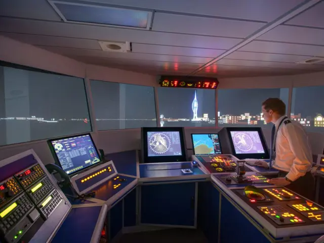 Illustrasjonsbildet fra innsiden av kommandobroen til et sjøfarende fartøy. Det er mørkt utenfor og flere instrumenter og skjermer lyser opp broen hvor en person står til høyre i bildet og hviler hendene på et tastatur. 