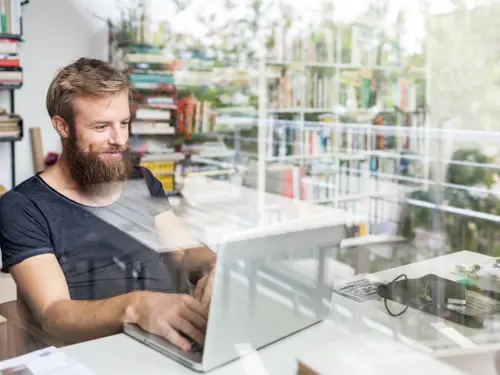 Ung mann med skjegg sitter foran laptop på hjemmekontor og smiler