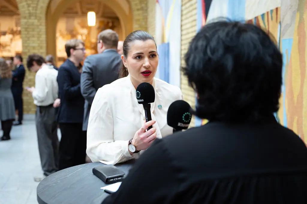 President Elisabet Haugsbø holder en mikrofon og snakker, hvit bluse, alvorlig uttrykk, folk i bakgrunnen i vandrehallen i Stortinget 