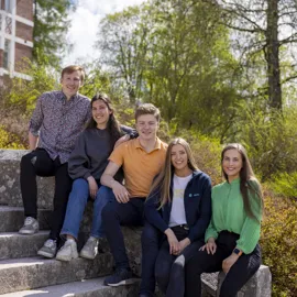 En rekke studenter som sitter i en trapp og smiler på en solfylt dag