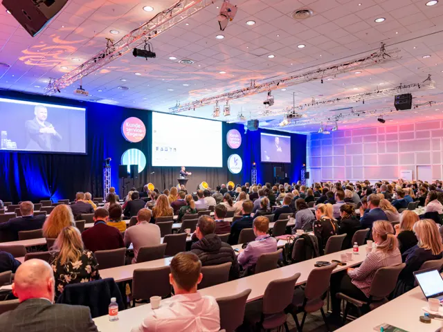 Oversiktsbilde av en scene og en sal med mange deltakere sittende ved bord