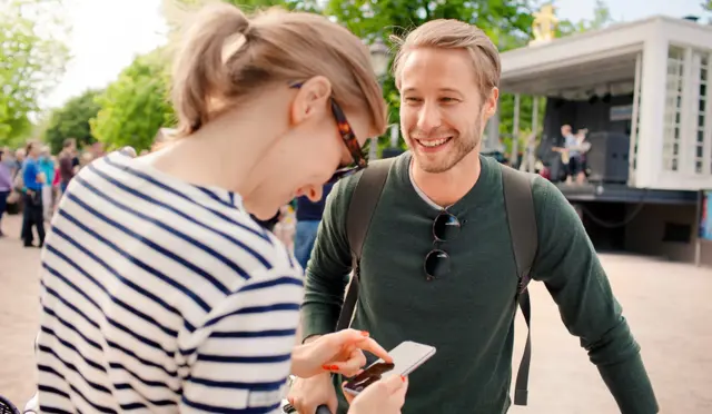 En ung mann smiler til en smilende kvinne med solbriller som har en telefon i hånda