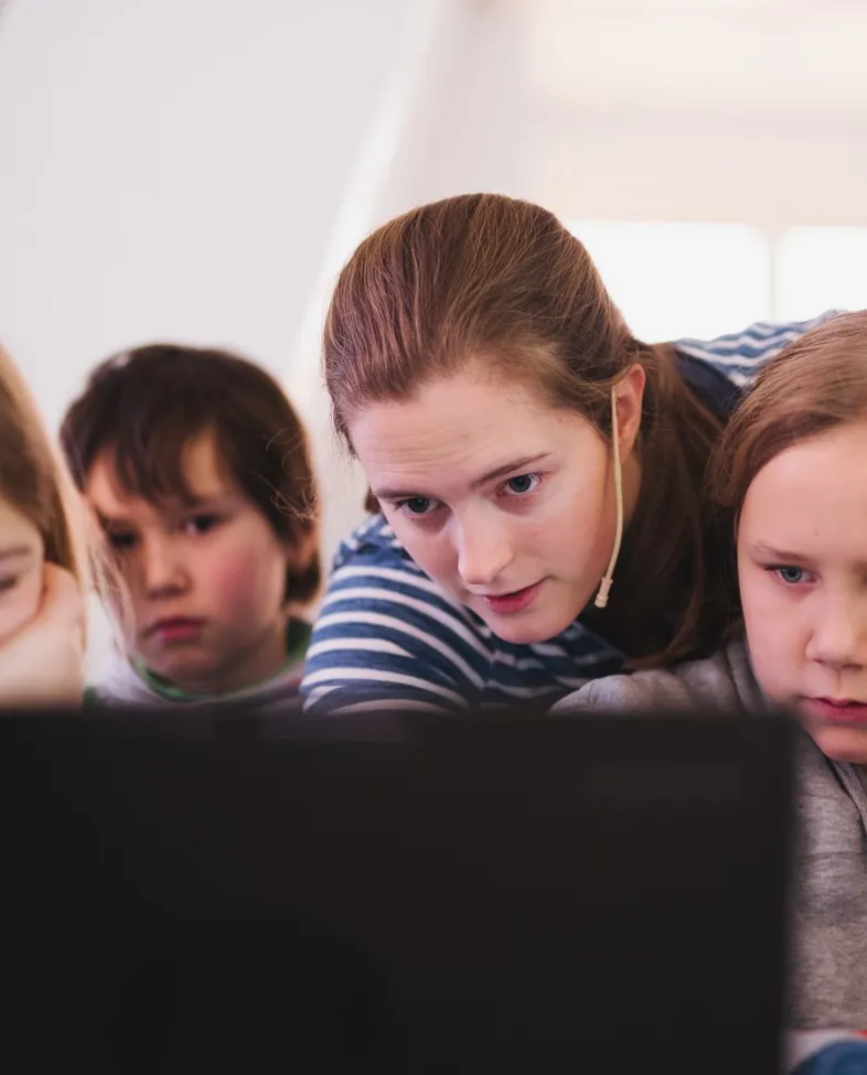En voksen kvinne peker på pc-skjerm i forgrunnen med tre barn rundt seg