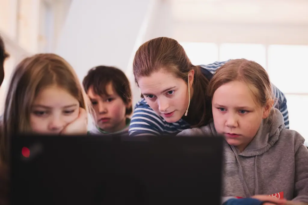En voksen kvinne peker på pc-skjerm i forgrunnen med tre barn rundt seg