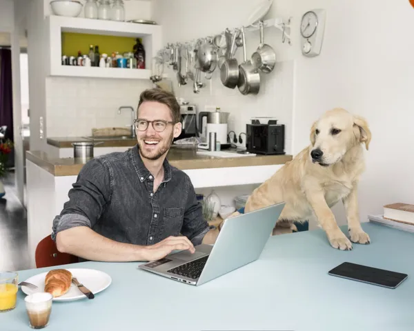 En ung mann på hjemmekontor ved kjøkkenbordet med en hund som sitter på stolen ved siden av han