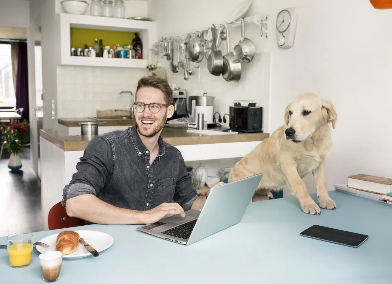 En ung mann på hjemmekontor ved kjøkkenbordet med en hund som sitter på stolen ved siden av han