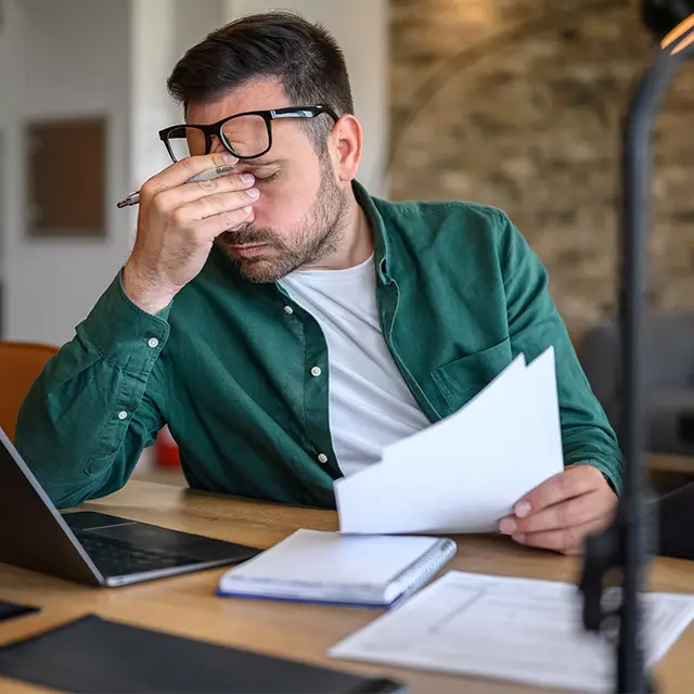 Mann ved skrivebord skyver brillen opp til pannen og holder seg i pannen