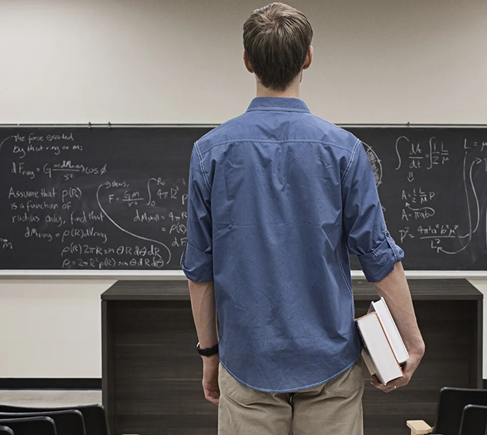 Mannlig student som står i en undervisningssal med to bøker under armen og ser mot tavlen