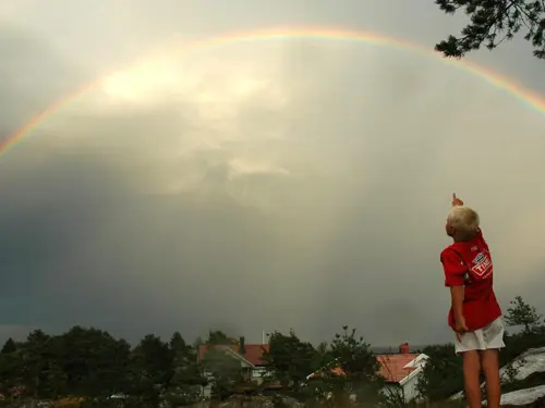 En ung gutt med blond hår peker opp til en stor regnbue