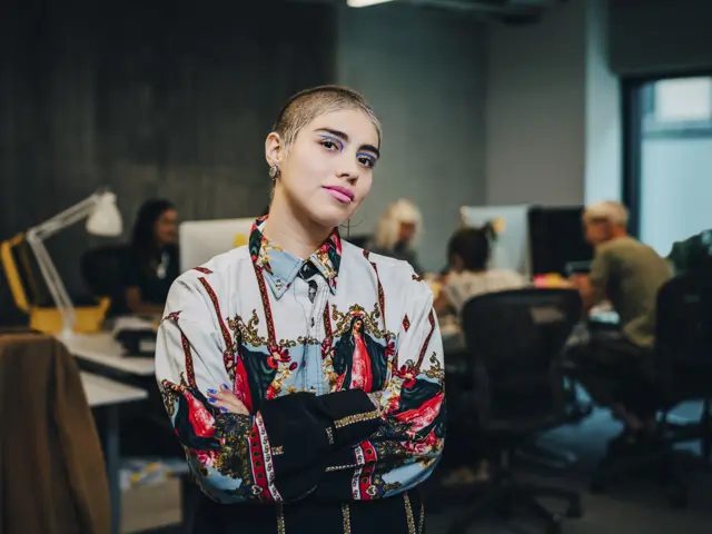 Kvinne med fargerik sminke og kort hår står i kontorlandskap med hendene i kors