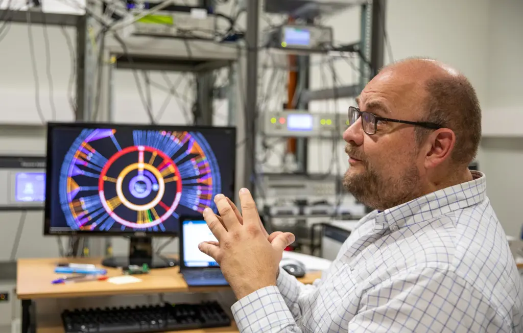 Harald Hauglin foran en dataskjerm som viser flere fargede sirkler utenfor hverandre - med enkelte små streker som binder sammen de ulike nivåene av sirkler. Harald gestikulerer med hendene. I bakgrunnen kan man ute av fokus skimte et stativ med flere utstyrsbokser med lysende knapper og skjermer og ledninger som går på kryss og tvers. 
