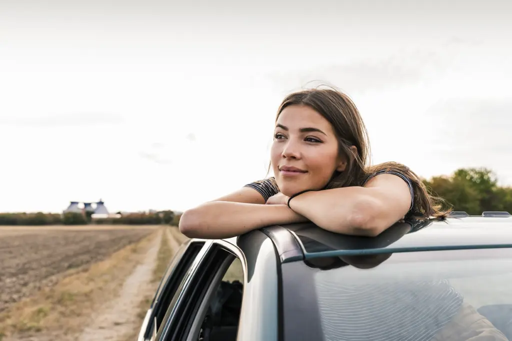 Kvinne som lener seg ut av solvinduet på en bil og ser ut i horisonten med et smil