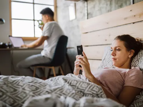 Working remotly, woman laying in bed on the phone, while a man is working on a computer besides the bed