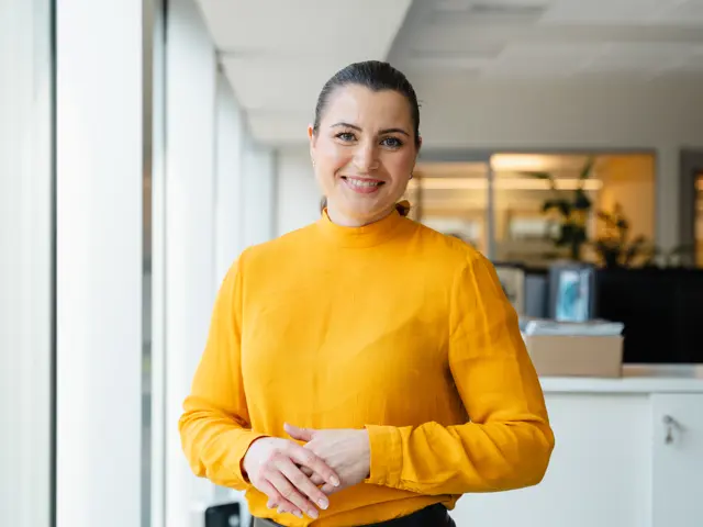 Elisabet Haugsbø smiler, med gul genser, i kontorlandskap