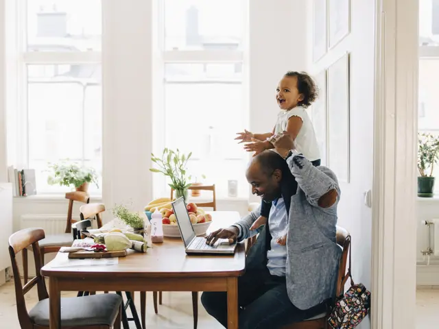 Picture of a Father working remotly from home the kichen table, with a child sitting on his back