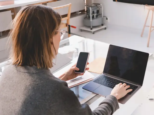 Person sitter foran en datamaskin med mobil i hånda