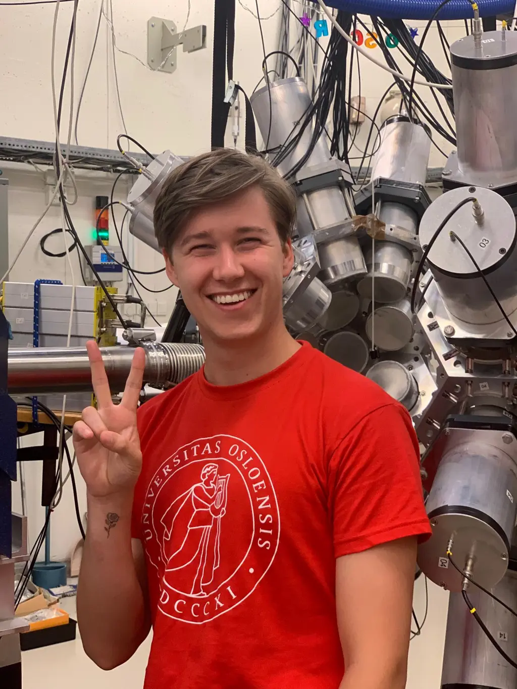 Foto av Pelle med rød UiO-tskjorte i en slags lab. Han holder opp to fingre og viser V-tegn. 