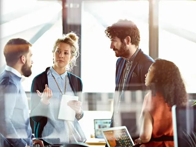 To menn og en kvinne lytter til kvinne som gestikulerer  i kontorlandskap