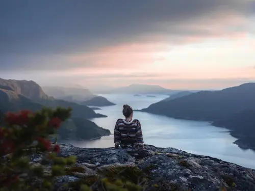 Illustrasjonsbilde av en ung kvinne i norgesgenser sittende på en fjelltopp med en fjord i bakgrunn