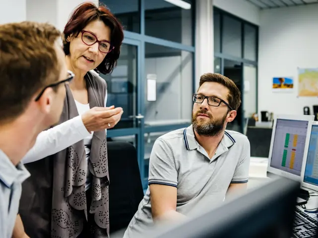 Illustrasjonsbilde av en kvinne som står og snatter til to unge menn som sitter ved datamaskiner