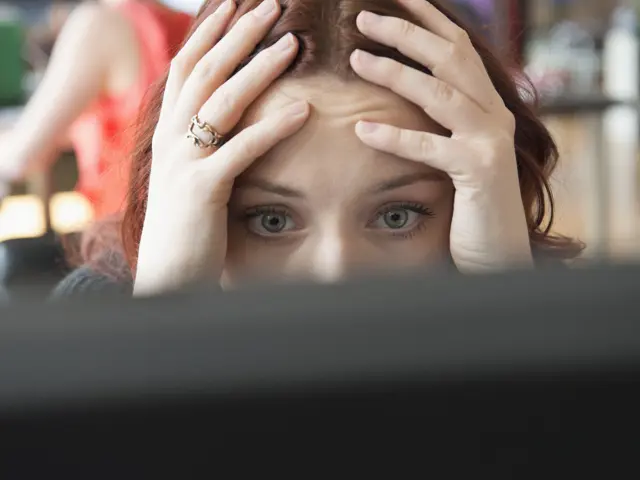 Close up of a woman in an office resting her head on her hands staring at a computer screen with an expressin of overwhelm