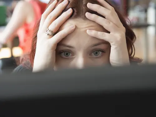 Close up of a woman in an office resting her head on her hands staring at a computer screen with an expressin of overwhelm