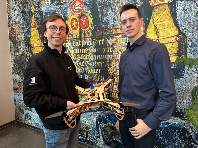 2 menn m drone foran vegg med kongesegl