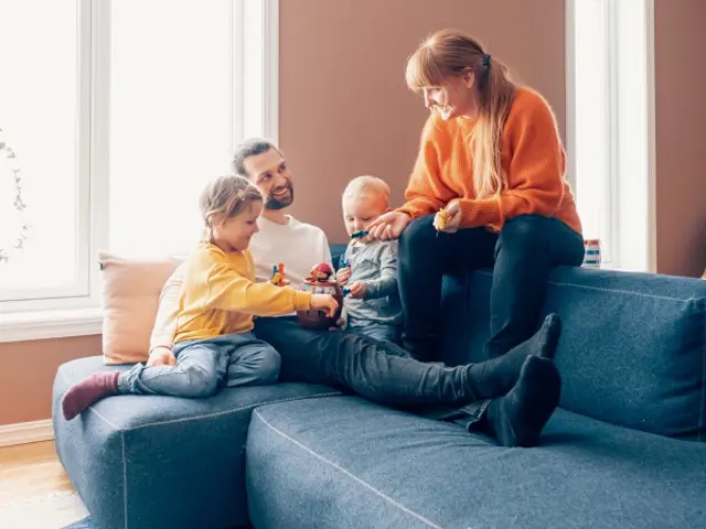 Bilde av en familie i en sofakrok. Mor, far og to barn som leker i fanget deres. 