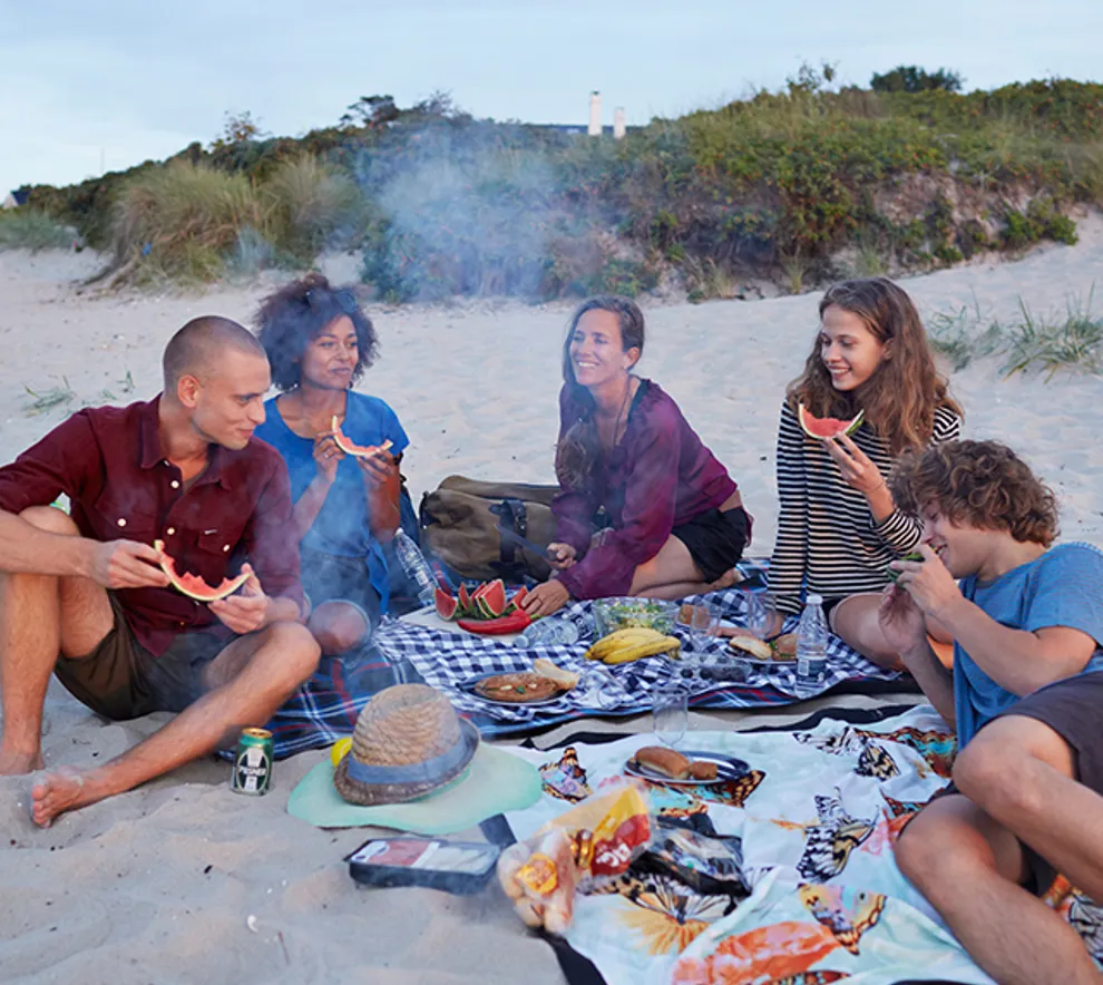 gruppe mennesker sitter på piknikkteppe på strand med grillmat