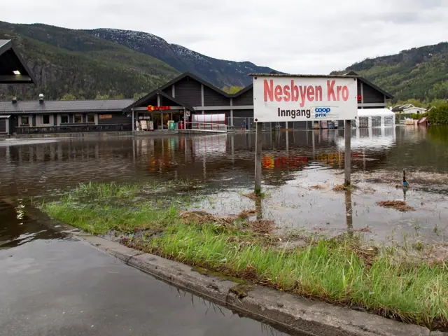 Skilt hvor det står "Nesbyen Kro - inngang Coop Prix" står i vann. Vannet går helt bort til bygninger man kan se i bakgrunnen. 