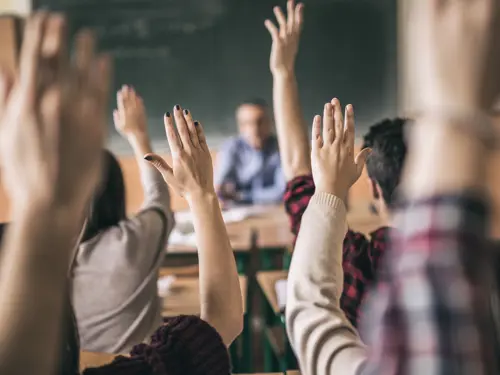 Klasserom med masse hender i været, i bakgrunnen en uklar lærer sittende ved en pult foran en grønn tavle 