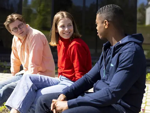 Tre studenter sitter i sola og snakker med hverandre