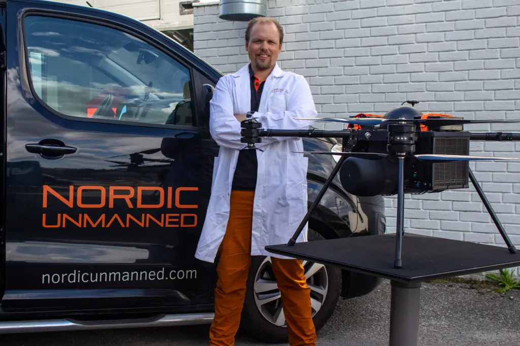 Jørgen Apeland i hvit labfrakk står ved siden av en drone og en bil