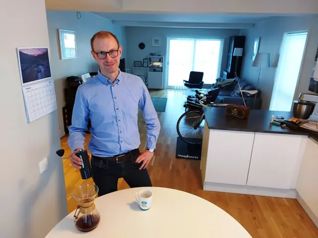 Grønmyr står ved kjøkkenbordet med kaffe i hånden og stuen i bakgrunn
