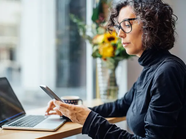 A woman with glasses typing on her phone while sitting at a laptop