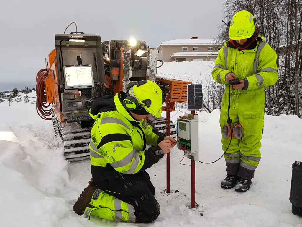 To personer i verneutstyr med måleutstyr og en gravemaskin i snølandskap