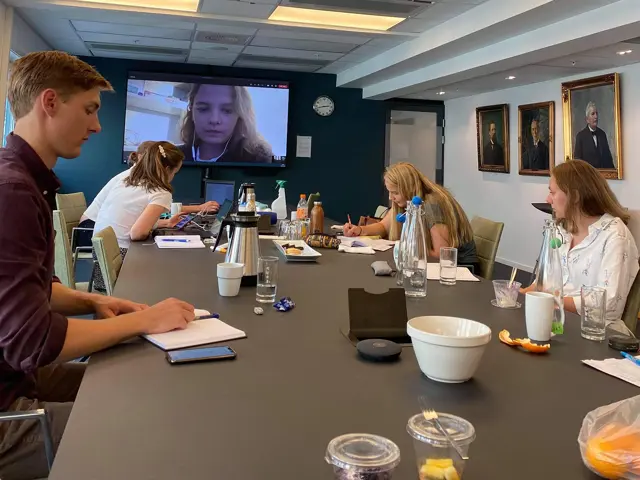 Et stort møterom med mennesker med 2m avstand, som ser på et videomøte på en stor TV-skjerm