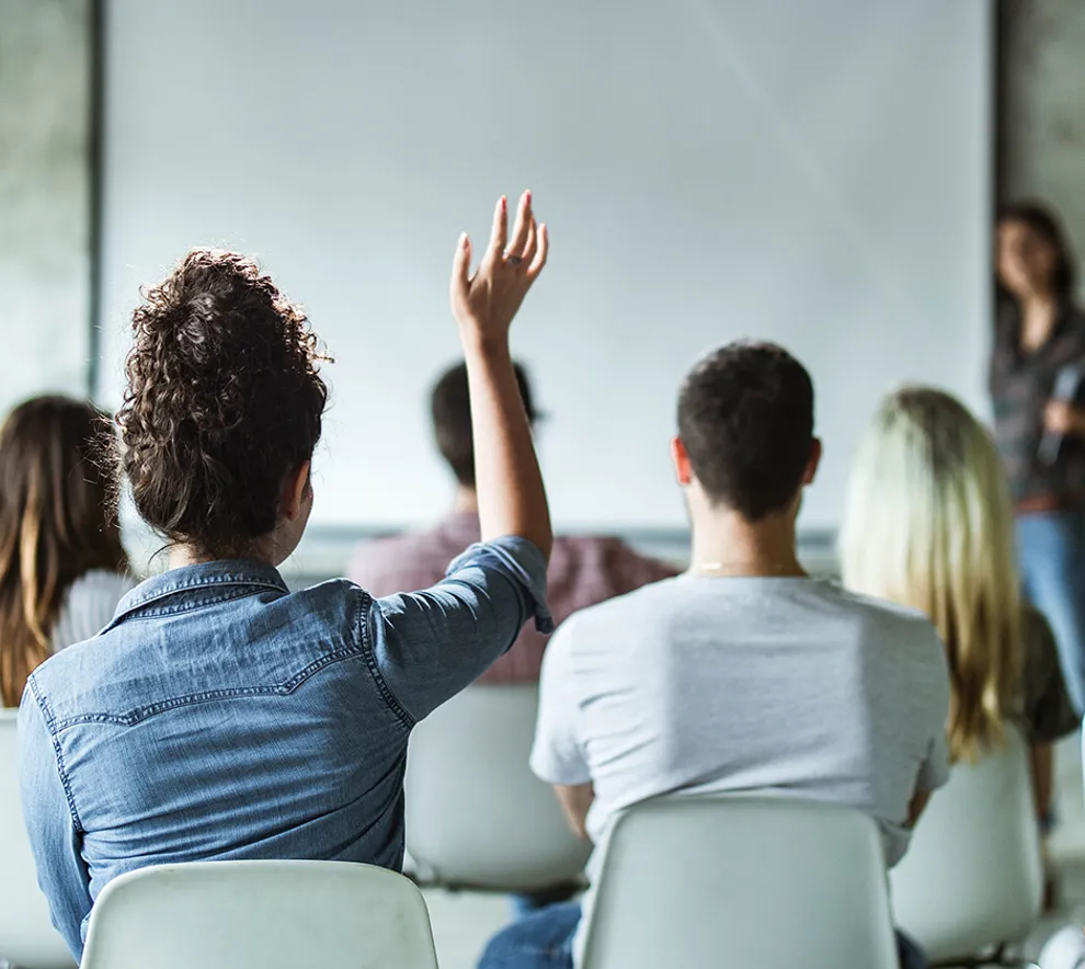 En forsamling mennesker som hører på en foredragsholder og en kvinne som rekker opp hånden