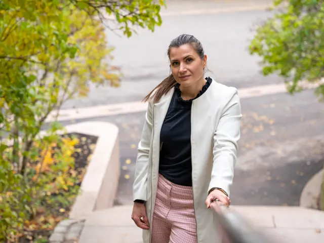 President Elisabet Haugsbø, hvit jakke, svart genser, rosa bukse, står utendørs nederst i en steintrapp med høyre hånd på rekkverket. Blader og trær rundt 