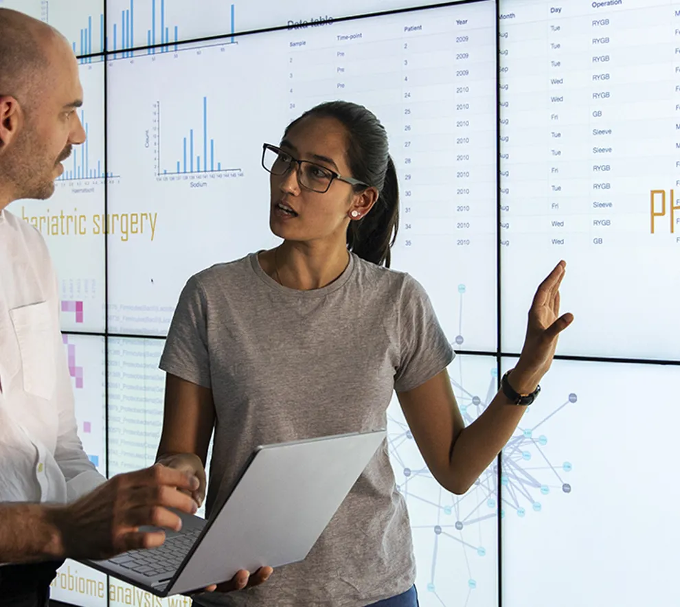 Kvinne og mann står ved stor skjerm som viser store datamengder.