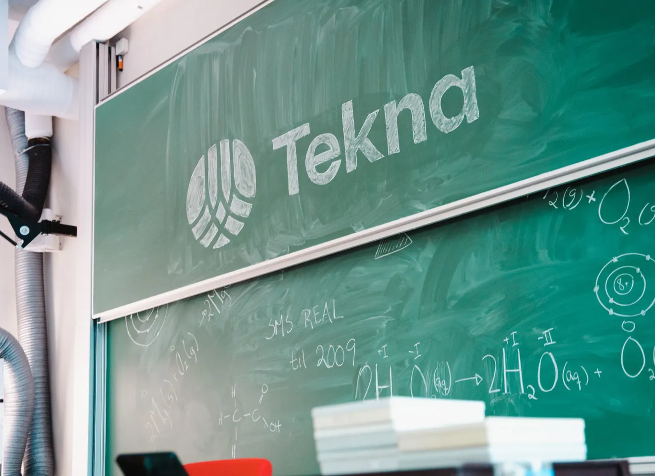Grønn tavle med formler og Tekna-logo skrevet i kritt