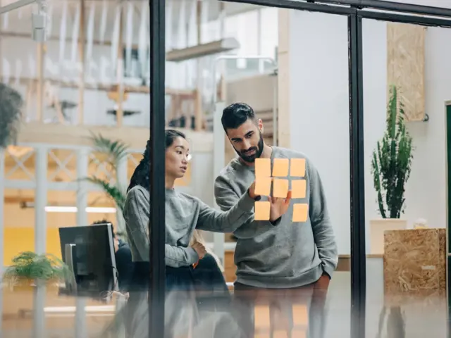 Mann og kvinne studerer post-it-lapper på en glassvegg