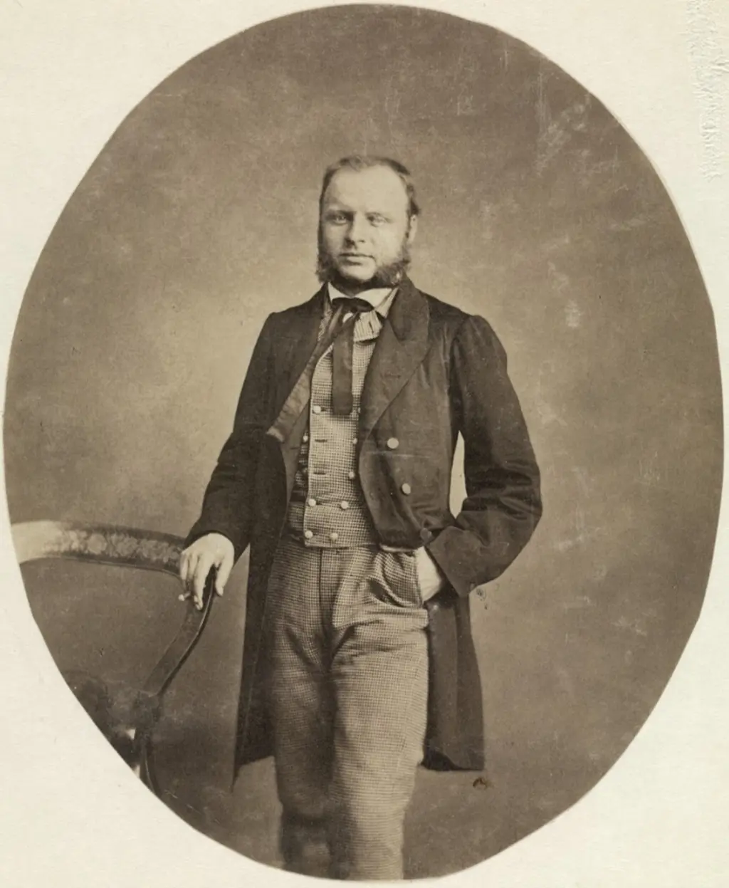 En stilig mann i en tredelt dress står ved siden av en stol. Han har venstre hånd på stolryggen og høyre hånd i lommen.